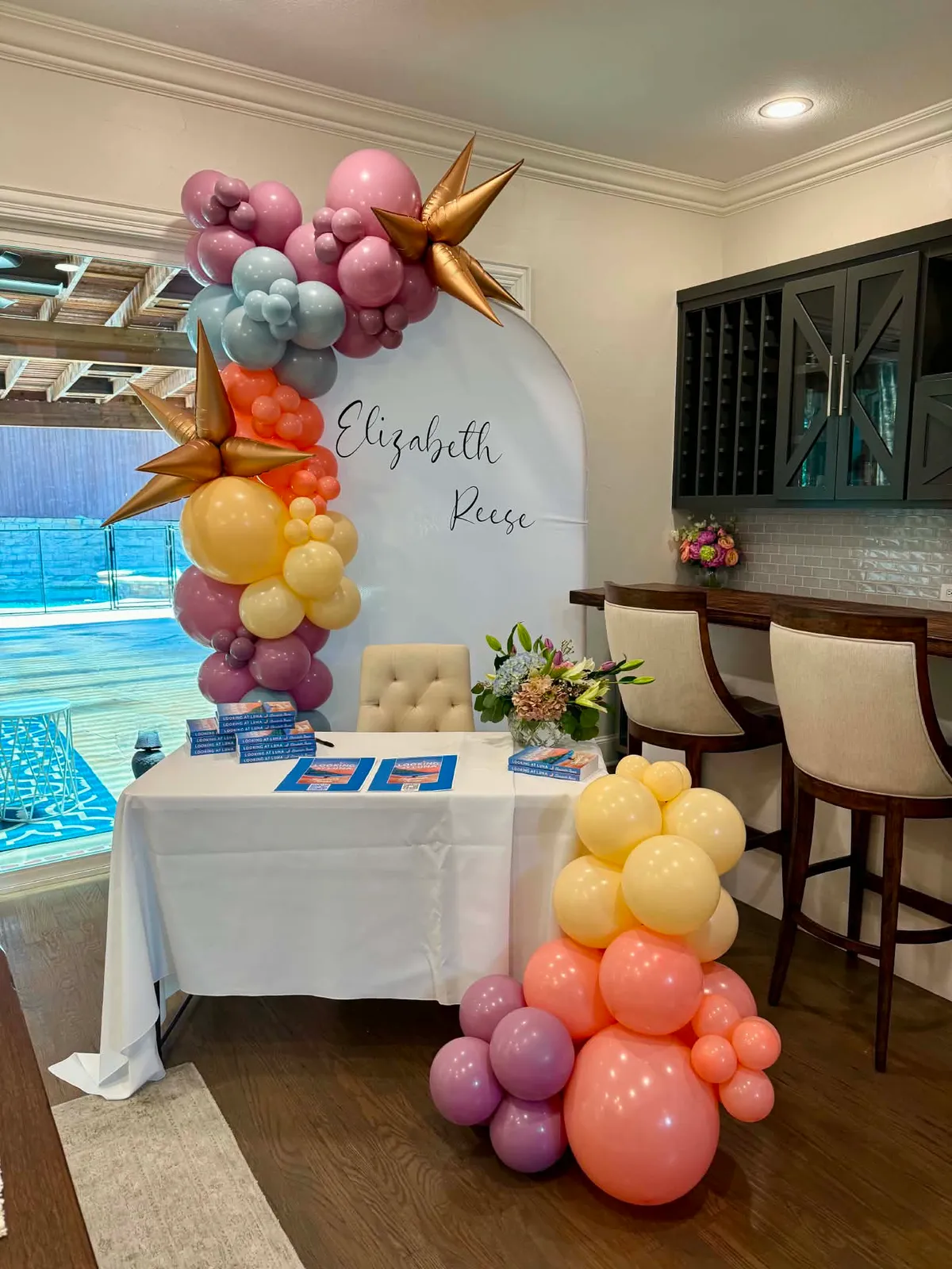 Pastel balloon garland in mauve, dusty blue, coral, and butter yellow with gold starbursts framing a white arch printed Elizabeth Reese above a book-signing table