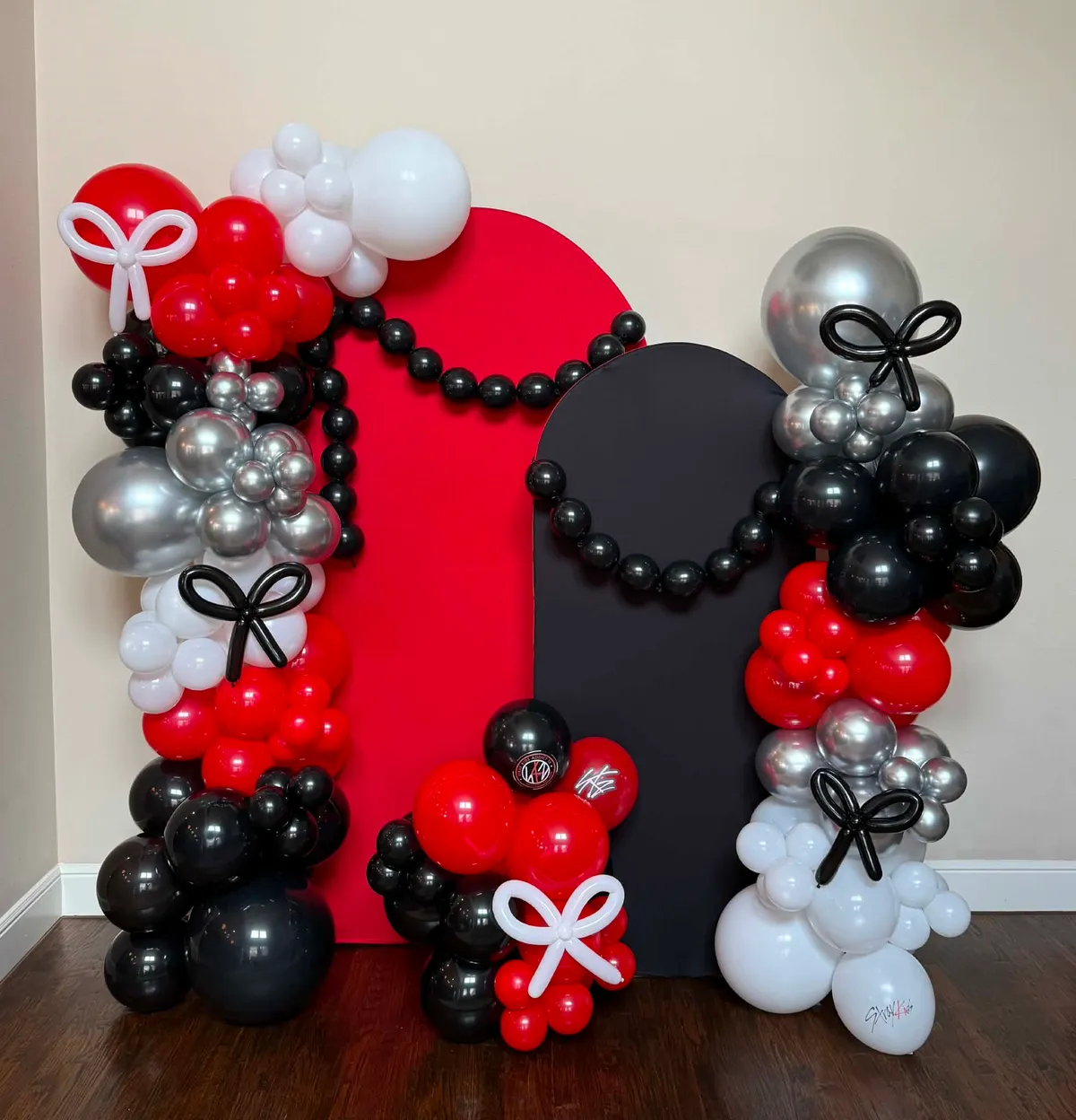 Red, black, white, and chrome-silver balloon setup with twin arches, black balloon-pearl swags, and balloon bows for a Stray Kids themed party