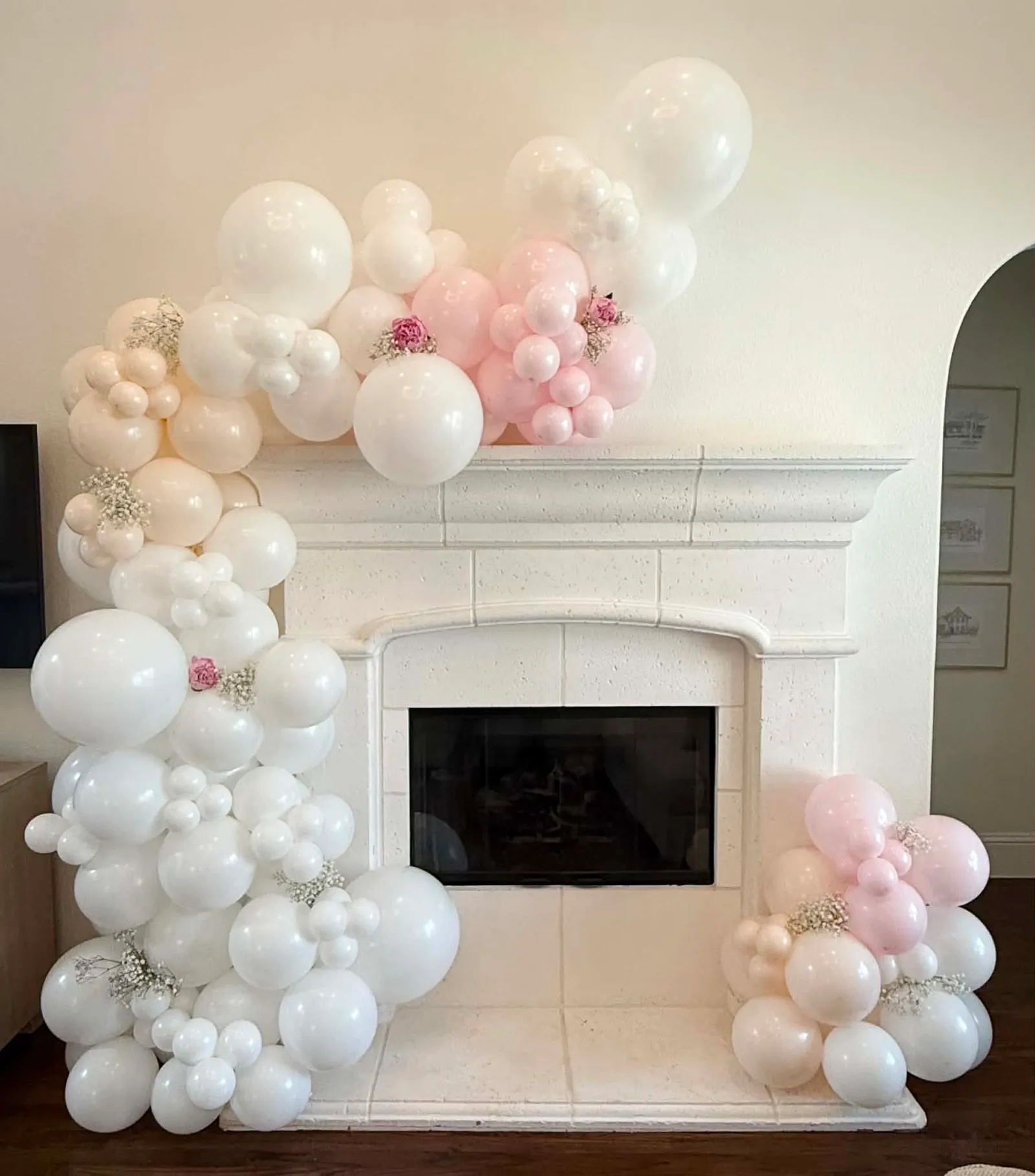 Soft white, cream, and blush balloon garland with dried baby’s breath and pink florals draped across a stone fireplace mantel