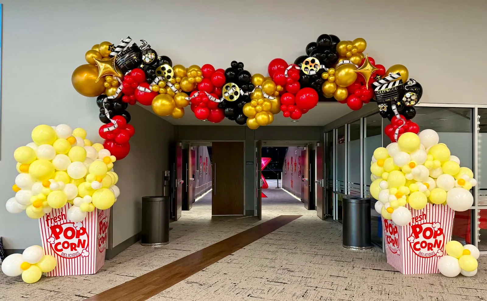 Wide view of a movie-night balloon garland in red, black, and gold arching over a doorway between two giant striped popcorn-bucket balloon columns